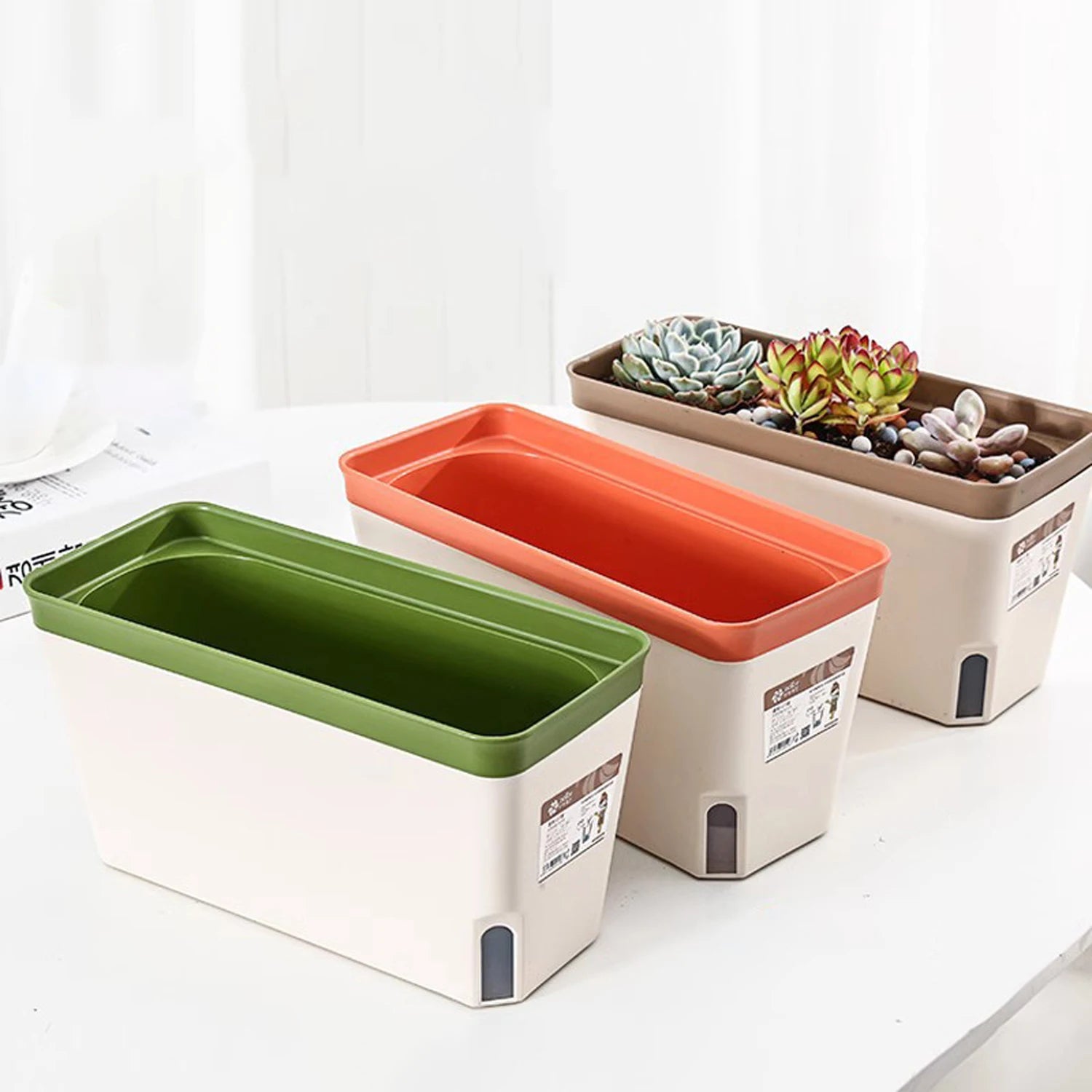 Rectangular planters in three colors, with one filled with succulents and another with empty pots, arranged on a table by a bright window.