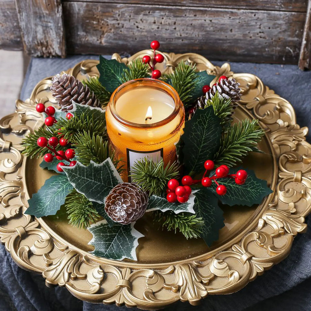 Christmas candle ring wreath featuring pine needles, holly leaves, pinecones, and red berries arranged on a gold tray