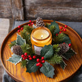 Artificial pine and holly candle ring wreath with red berries and pinecones styled around an amber jar candle on a wood tray