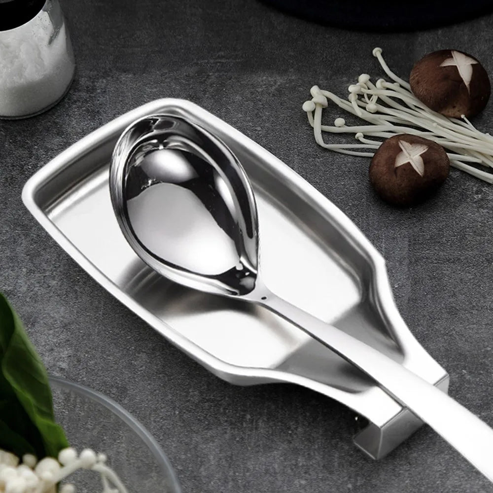 Stainless steel spoon rest holding a serving spoon on a kitchen counter with fresh vegetables and mushrooms nearby