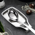 Stainless steel spoon rest holding a serving spoon on a kitchen counter with fresh vegetables and mushrooms nearby