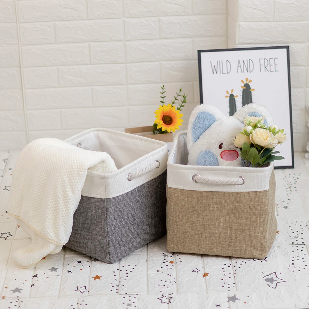 Two collapsible fabric storage cubes with rope handles holding a blanket and toys, styled on a white floor in a cozy home setting