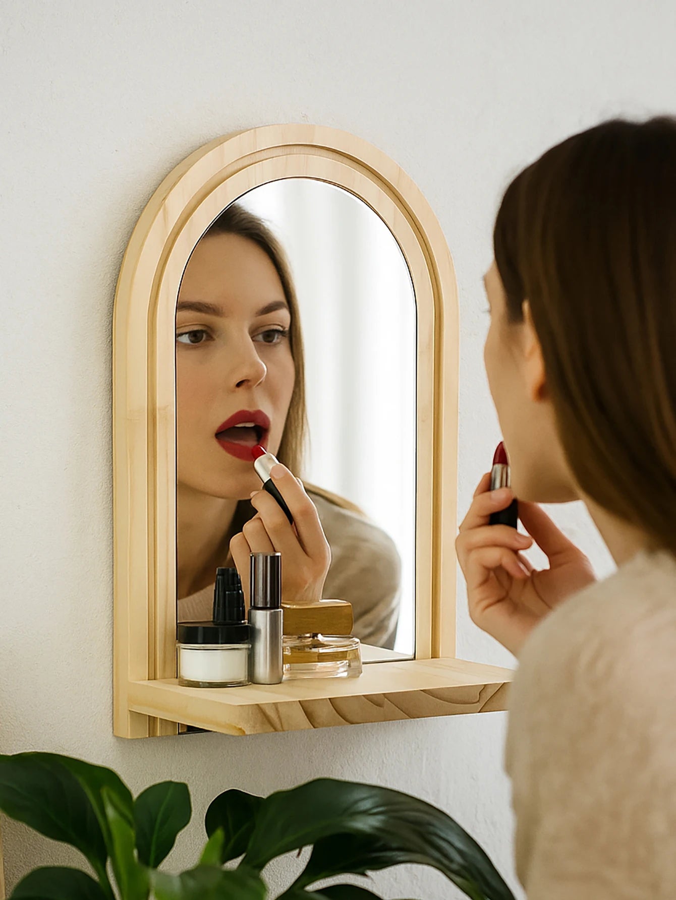 natural wood arched wall mirror with shelf in use as a vanity, with a person applying lipstick and makeup jars on the shelf