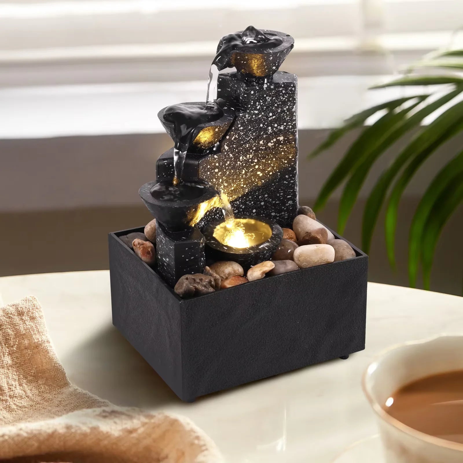 Three-tier LED tabletop water fountain with natural pebbles on a table, water flowing with warm light glow
