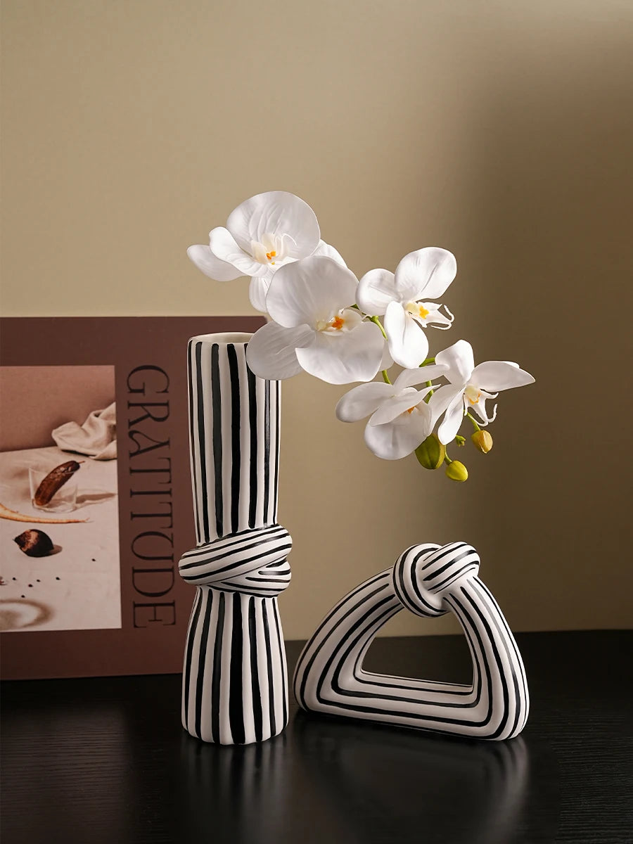 Two black and white striped ceramic vases with knot designs on a table, one tall and cylindrical, the other triangular and sculptural, both styled with white orchid flowers.