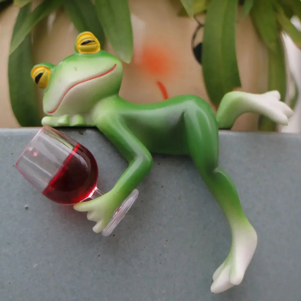 Close-up of a resin frog decor figure reclining on a planter edge and holding a glass with red liquid