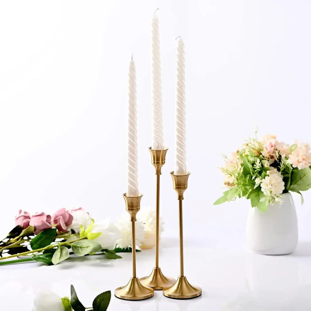 Trio of slim, gold metal candle holders with white twisted taper candles, surrounded by floral arrangements on a white background.