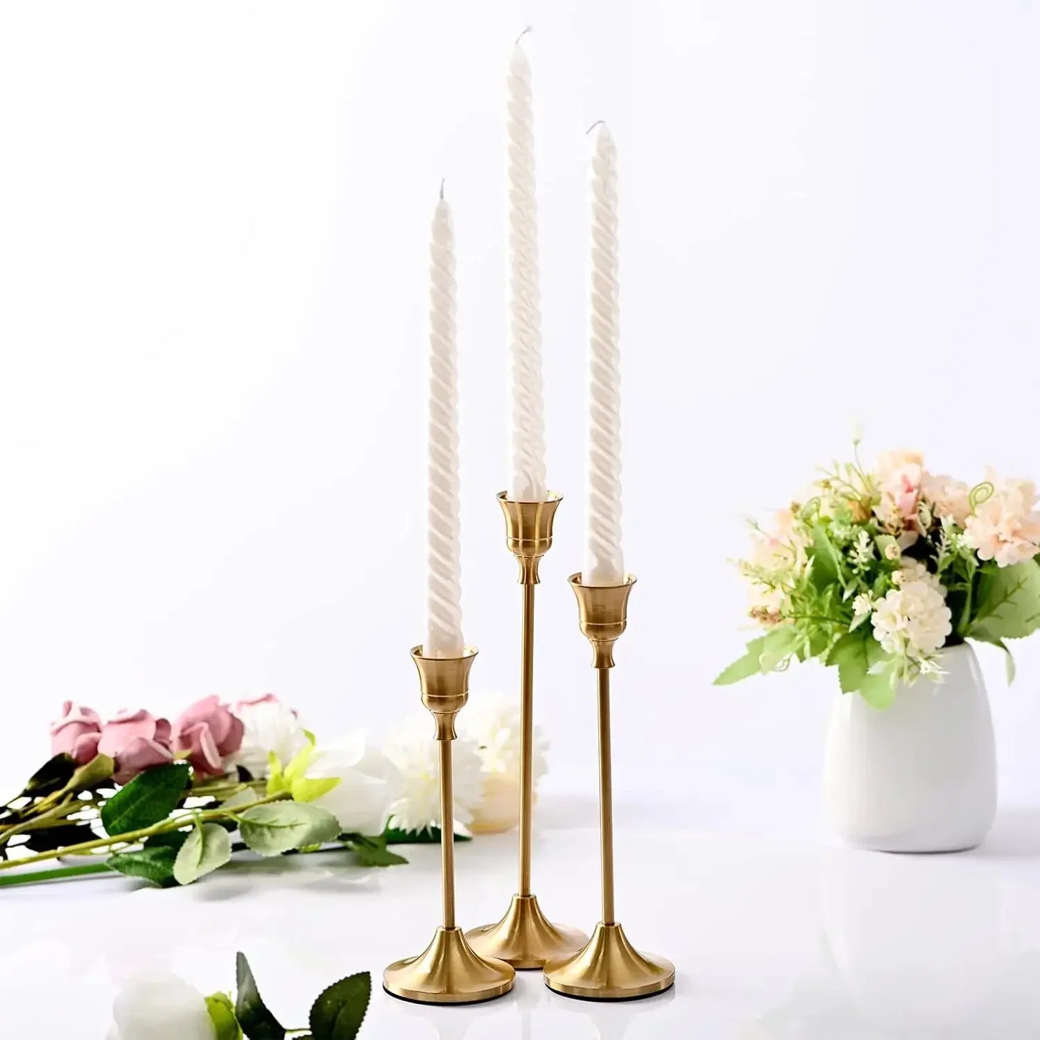 Trio of slim, gold metal candle holders with white twisted taper candles, surrounded by floral arrangements on a white background.