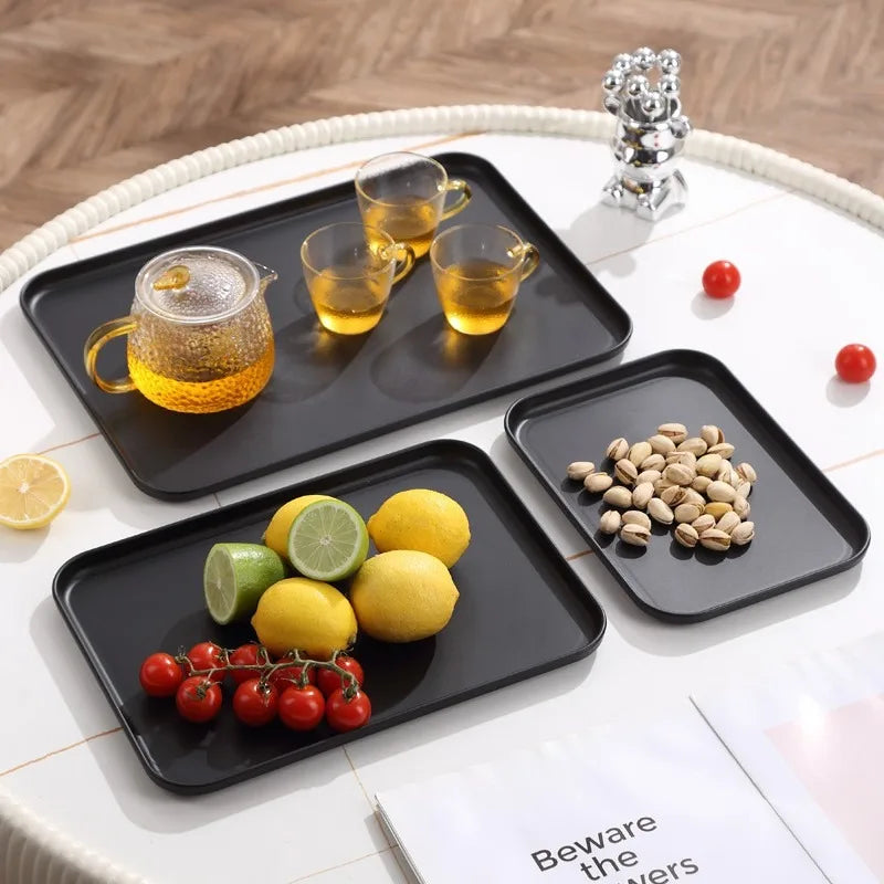 Black rectangular trays on a white table, one holding a glass teapot and cups, one with lemons and limes, one with pistachios