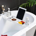 White expandable bathtub tray holding tablet, soap dispenser, glasses, and snacks on a modern tub