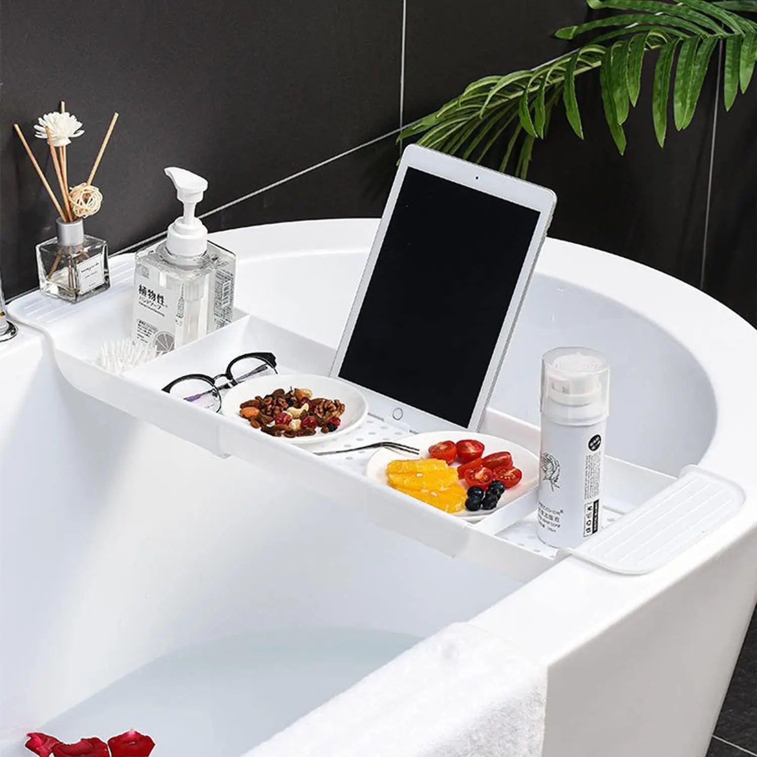 White expandable bathtub tray holding tablet, soap dispenser, glasses, and snacks on a modern tub