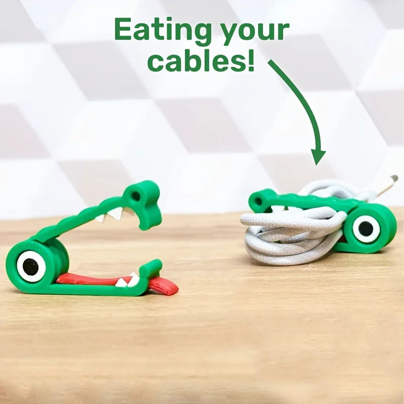 Two green crocodile-shaped cable organizer clips on a wood surface, one holding a coiled charging cable