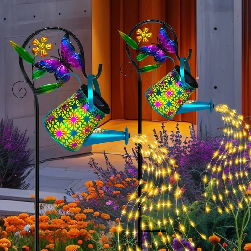 Two solar butterfly watering can lanterns lighting a garden path at dusk, showing the gentle warm light effect on flowers.