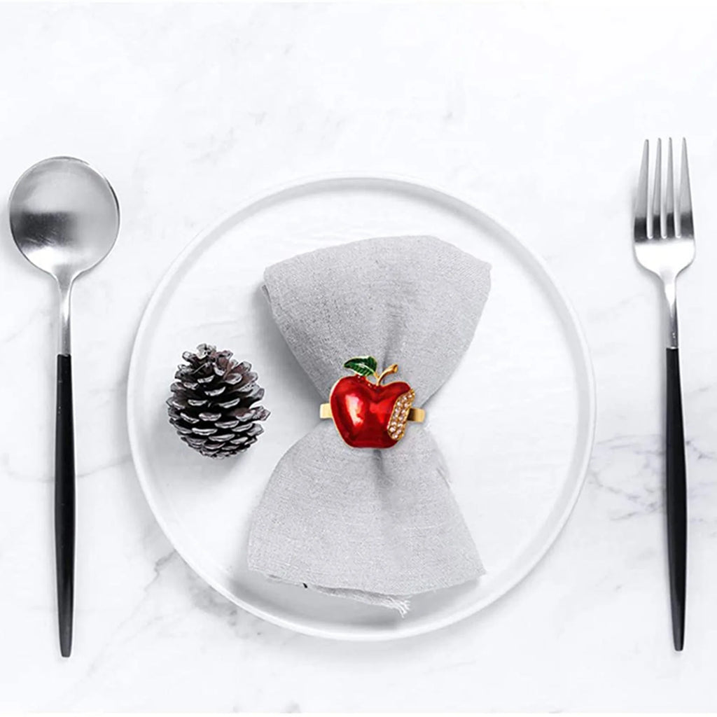 Apple napkin ring securing a grey cloth napkin on a white plate, with pinecone and cutlery nearby