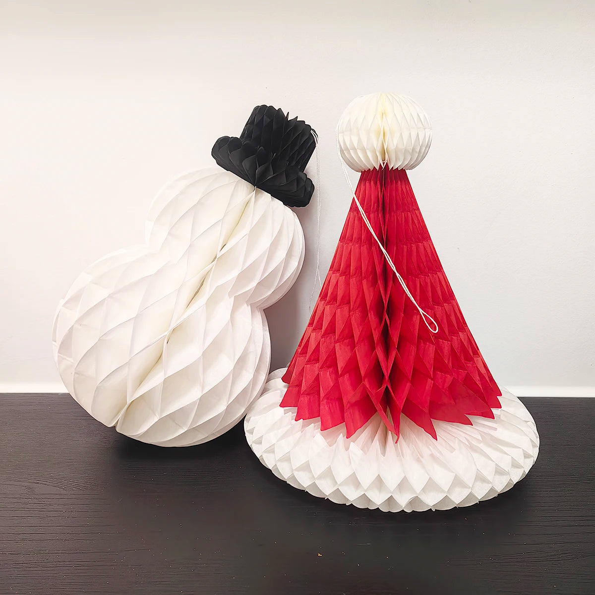 Honeycomb paper snowman and red Santa hat ornaments displayed side by side on a black surface