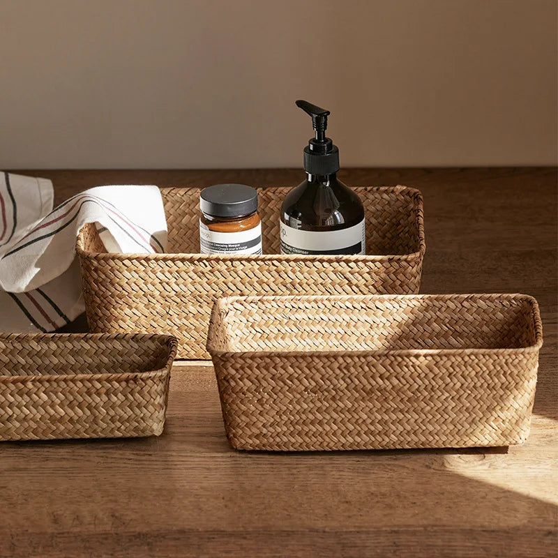 Three handwoven rectangular seagrass storage baskets on a wooden surface, holding toiletries and a cloth
