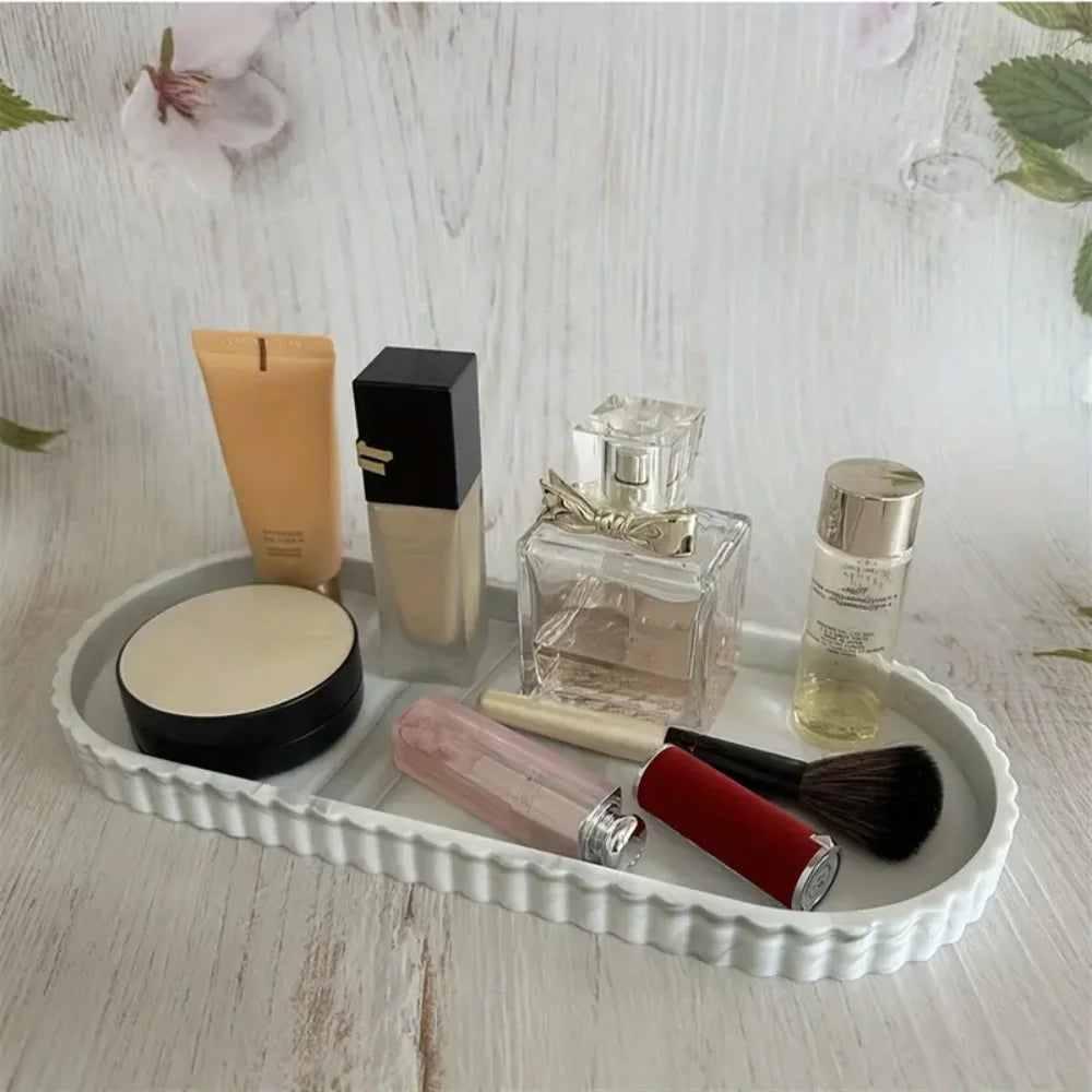 White ridged oval silicone tray holding perfume, makeup, and a makeup brush on a light wooden counter