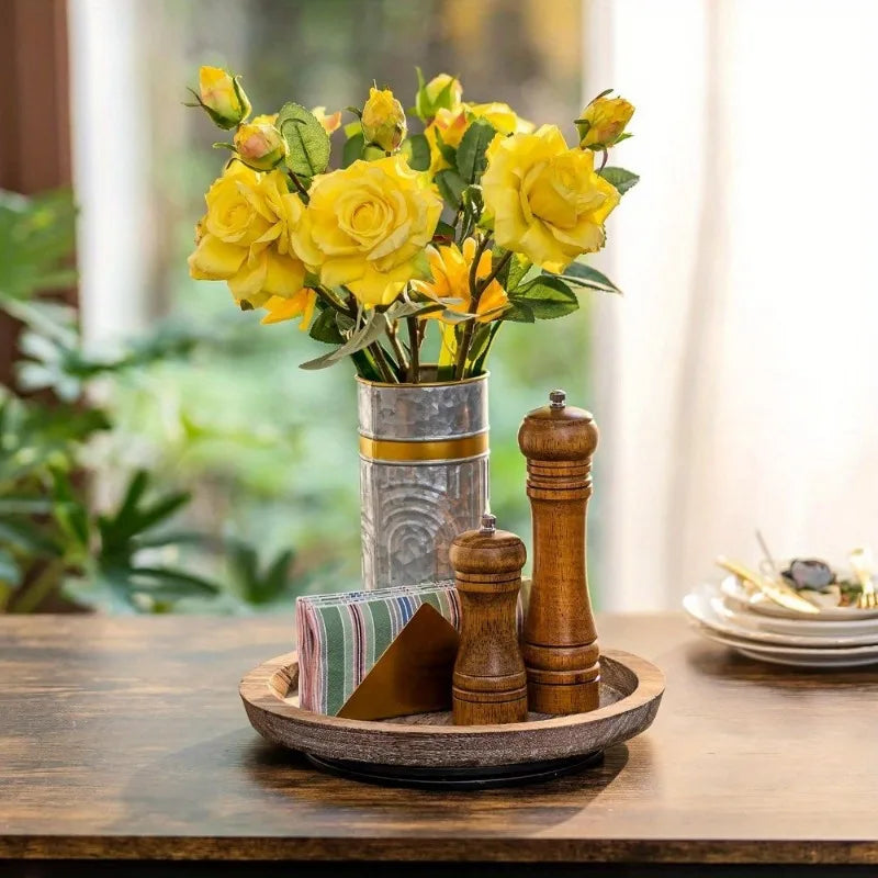 Round wooden tray on a dining table holding a vase of yellow flowers, pepper grinders, and napkins