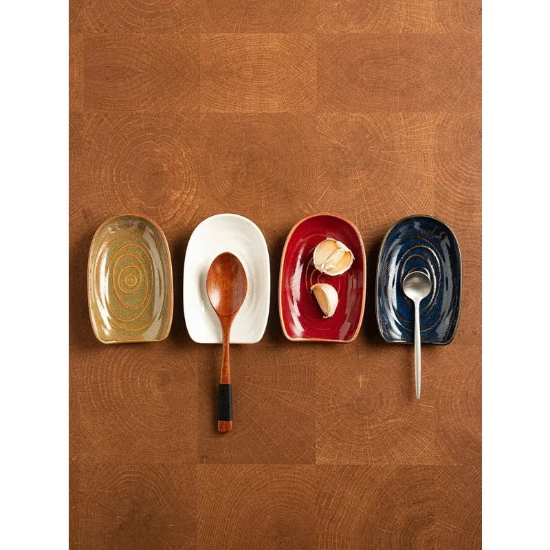 Four oval ceramic spoon rests in brown, white, red, and blue, each with a spoon or garlic shell on a wooden countertop