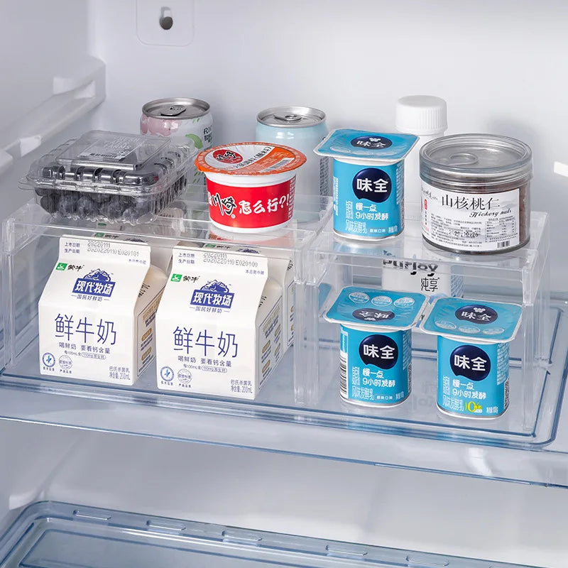 Clear stackable cabinet risers holding milk cartons, yogurt, and cans in a refrigerator