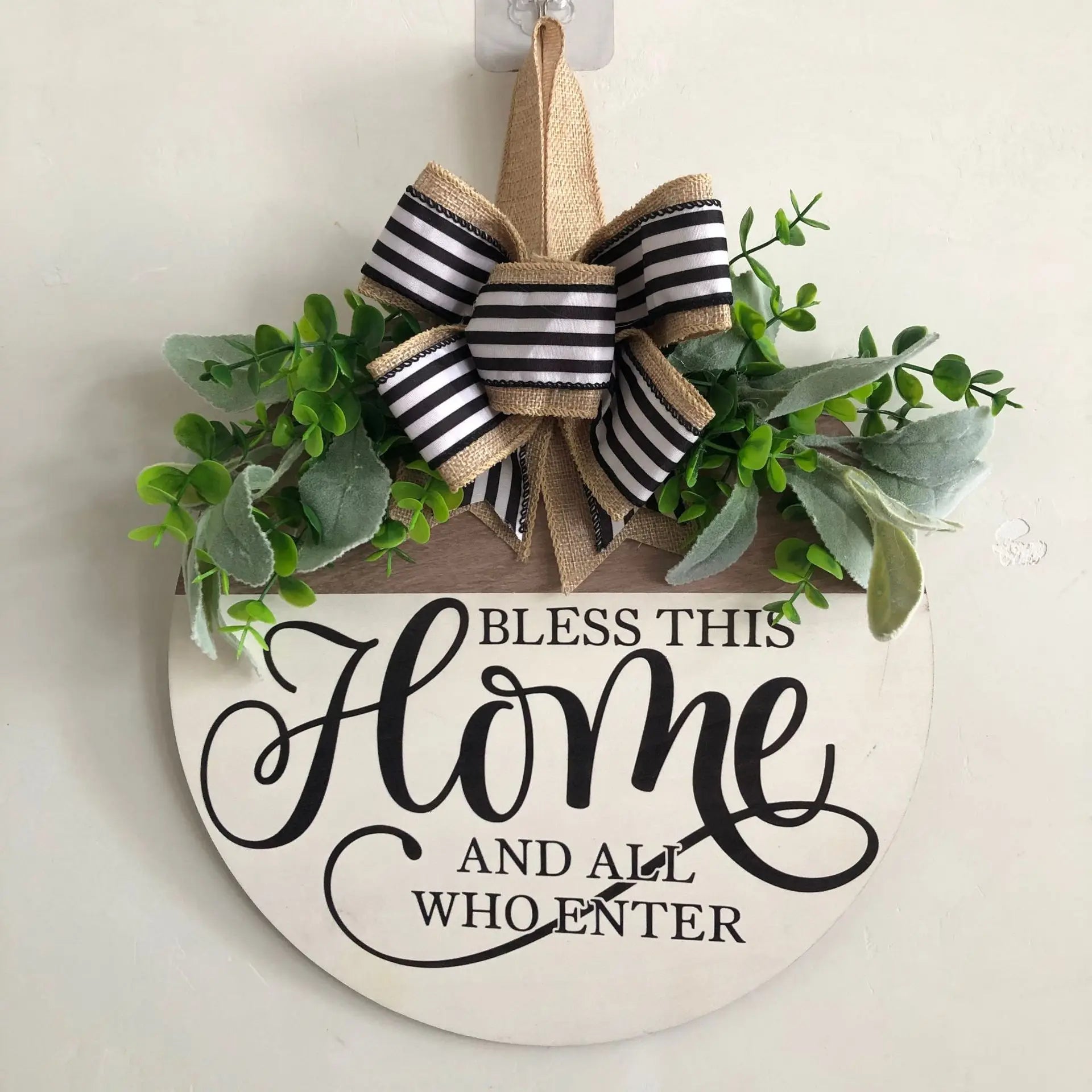 Round wooden welcome wreath sign with black and tan striped bow, faux greenery, and 'Bless This Home and All Who Enter' text