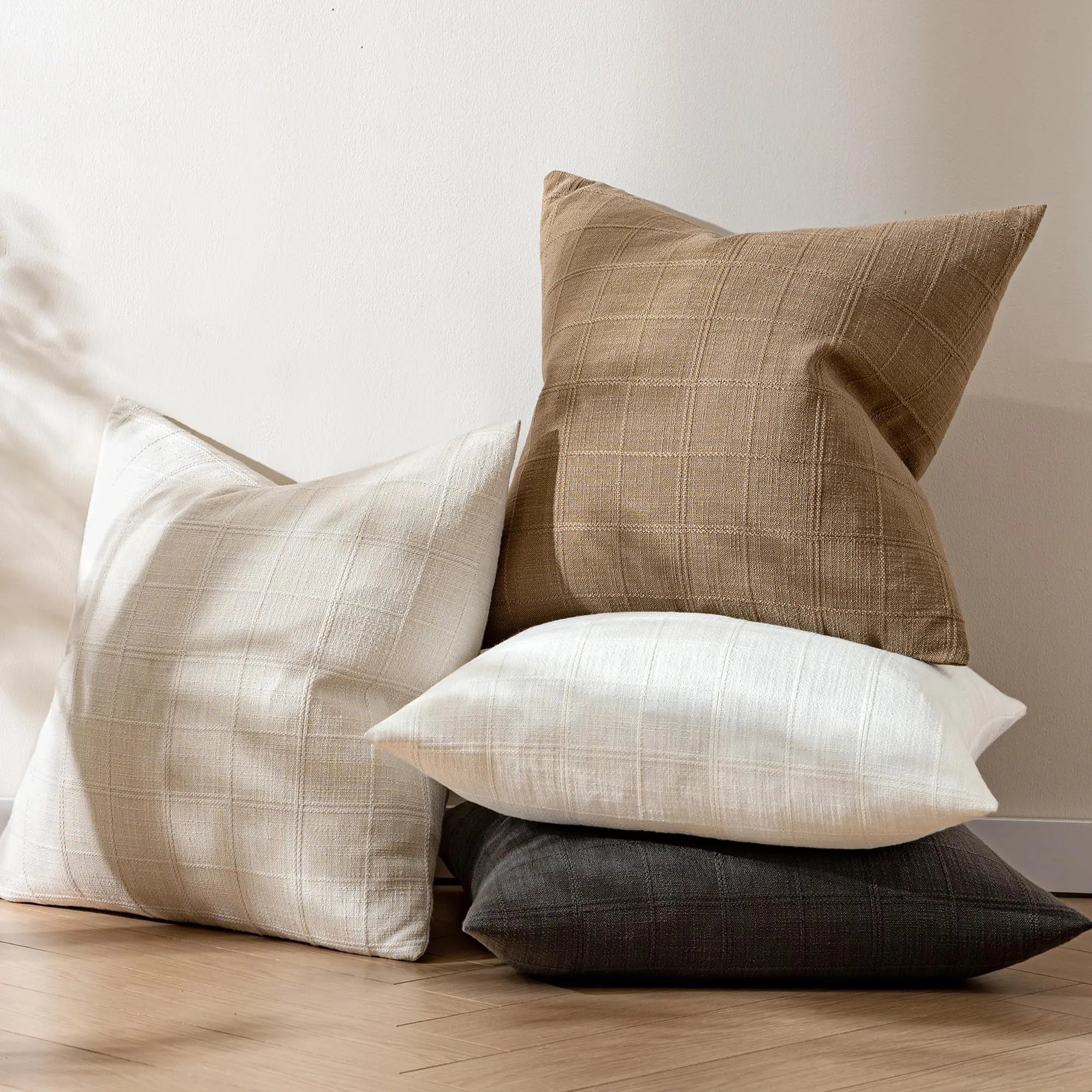 Neutral plaid pillow covers in beige, brown, white, and black stacked casually on a wooden floor