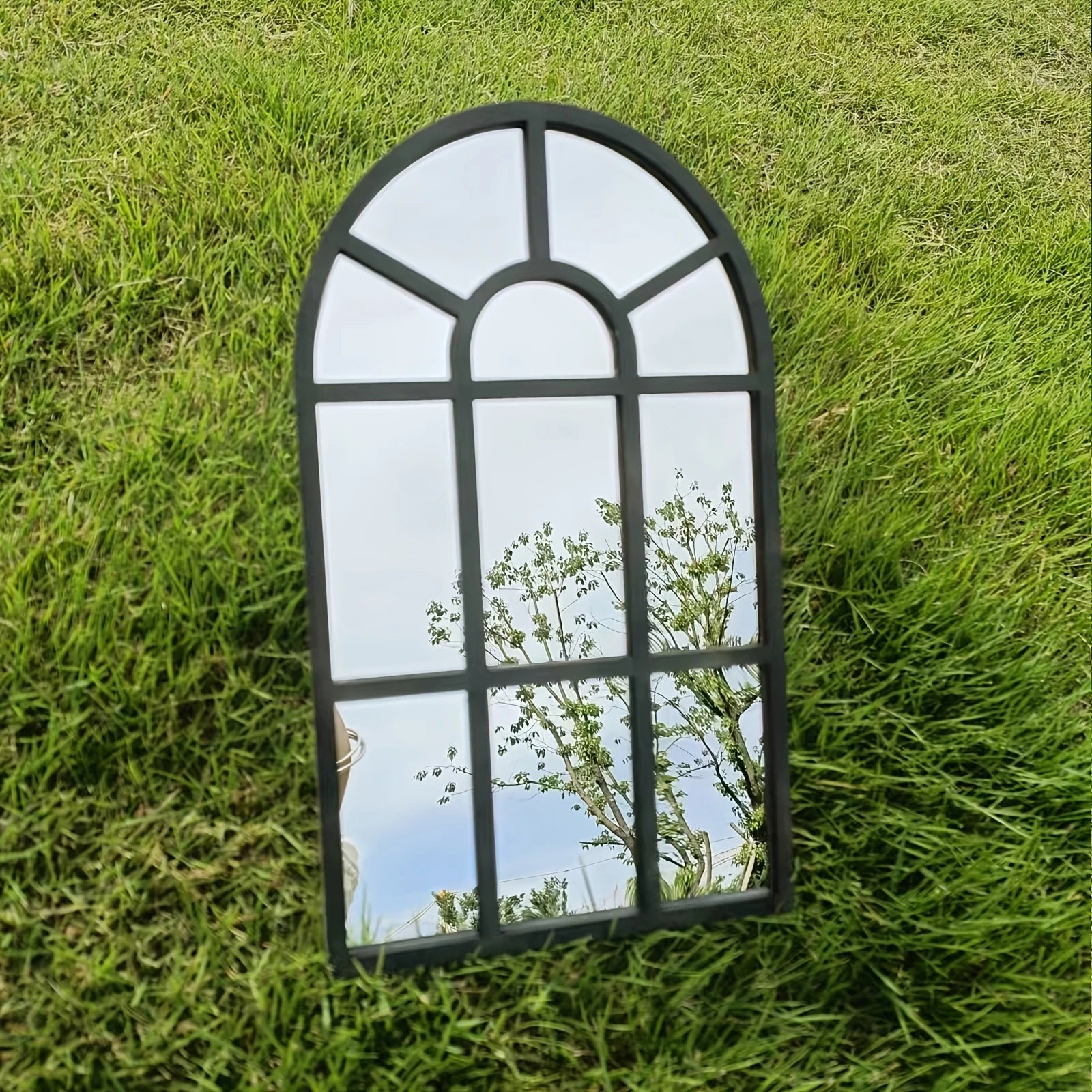 Gothic arch black windowpane wall mirror displayed outdoors on grass reflecting sky and trees