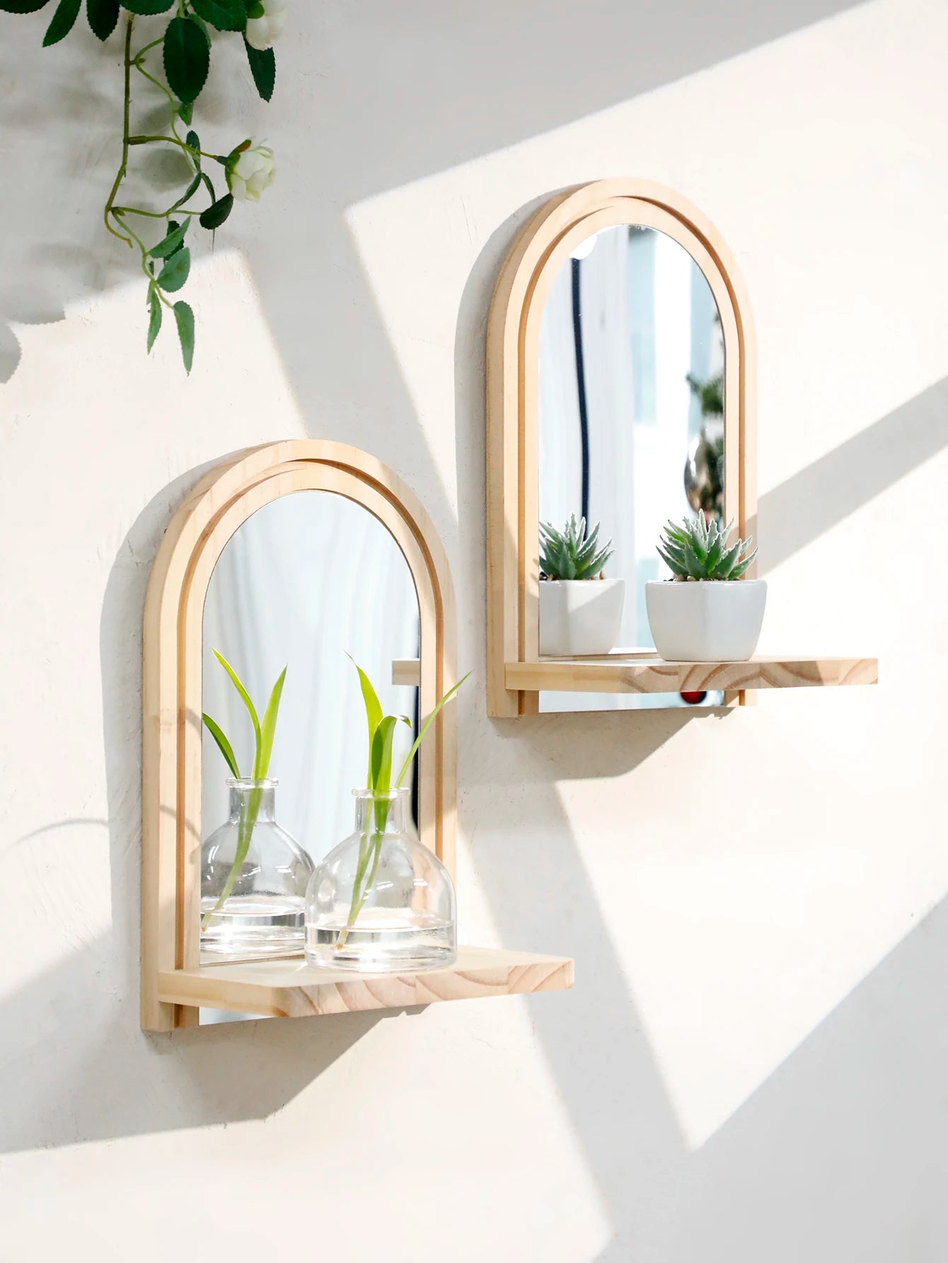 two arched wall mirrors with shelves, each holding small plants and glass vases, mounted on a white wall