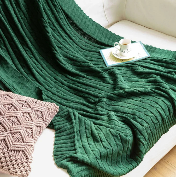 Green cable knit cotton throw blanket draped over a sofa with a cup and book