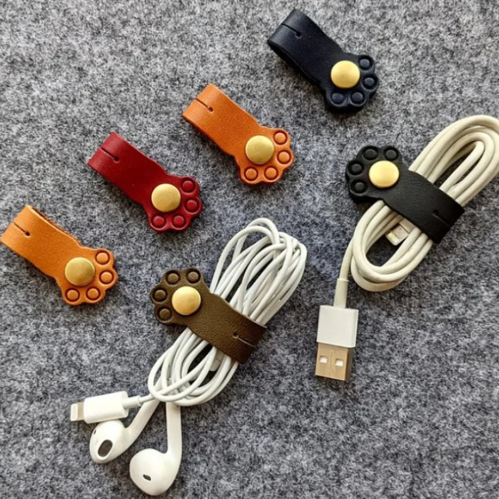 Cat paw shaped leather cable organizers in four colors shown securing earphone and charging cables on a felt surface