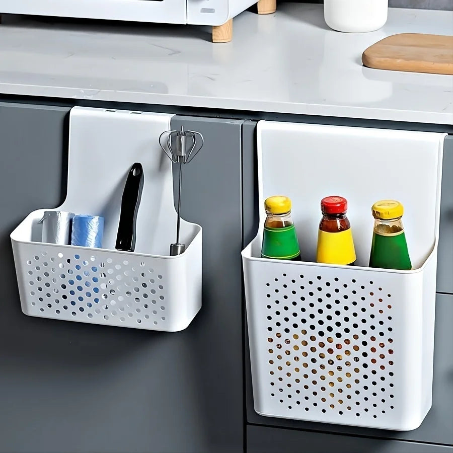 Two white plastic over-cabinet storage baskets hanging on dark kitchen cabinet doors, one holding small kitchen tools and cloths, the other holding condiment bottles.