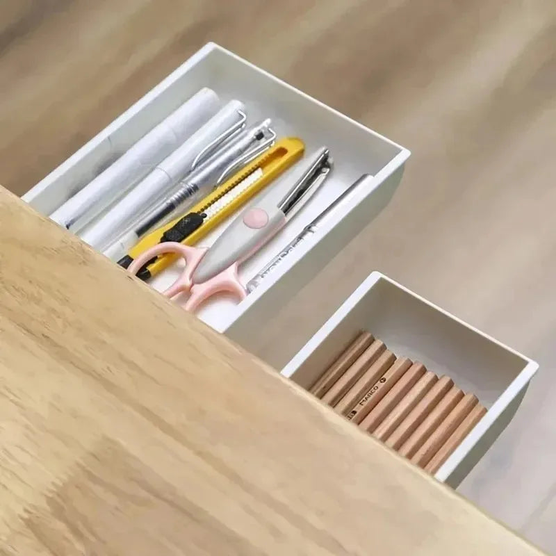 White plastic under-desk storage drawers organizing pens, scissors, and pencils under wooden table