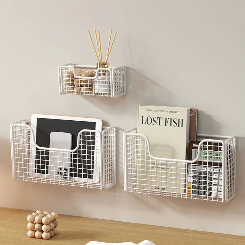 Three white iron storage baskets mounted on a wall, holding books, a tablet, and decor items