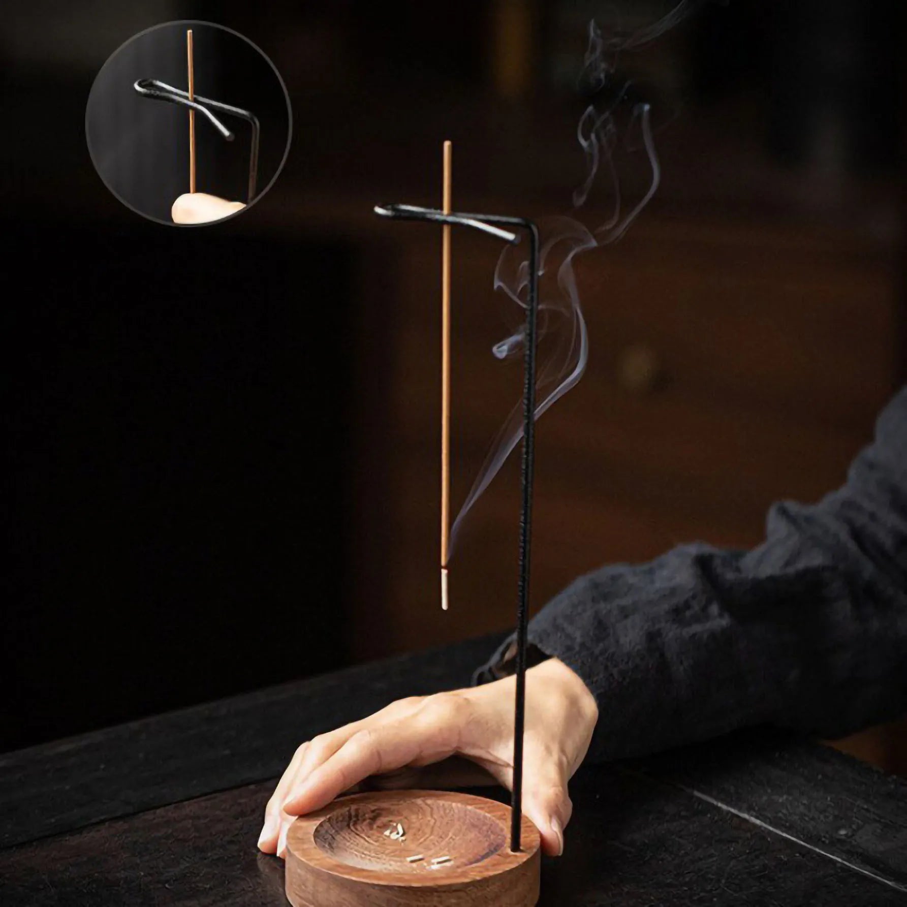 Hand holding the base of a wooden incense holder with metal arm suspending a burning incense stick, showing ash collected in tray