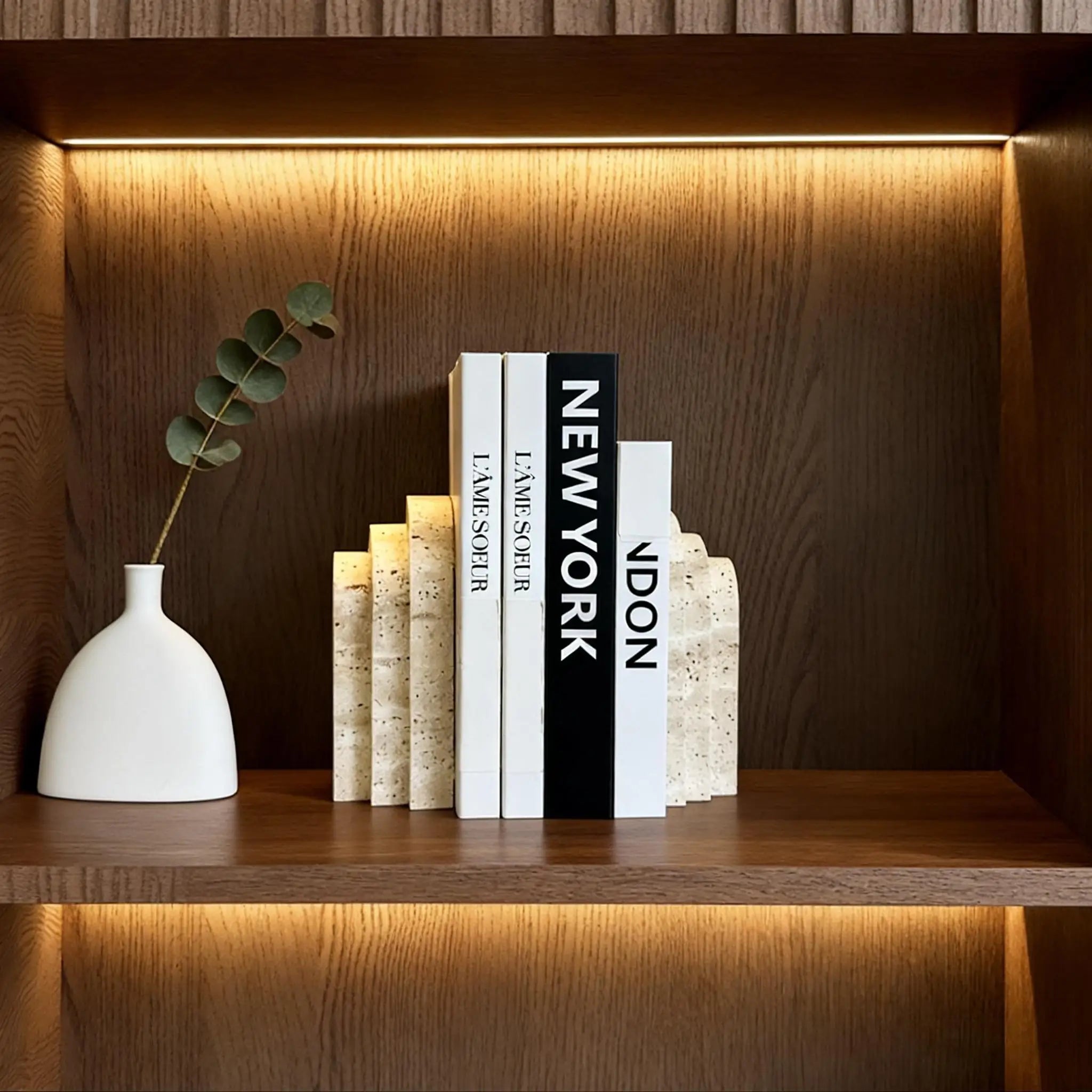 Travertine marble bookend supporting books on a wooden shelf, accented by a white vase and warm lighting