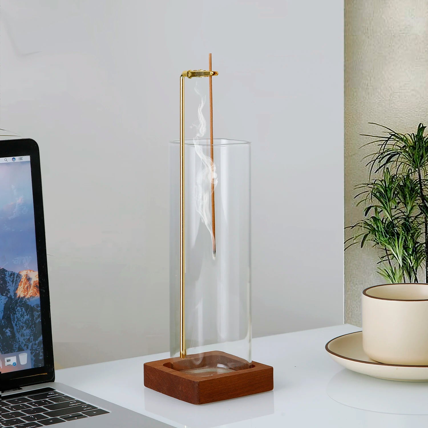 Glass incense holder with brass arm and wooden base placed beside a laptop and cup