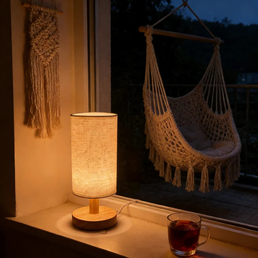 Wooden bedside lamp with a linen shade on a windowsill next to a macrame wall hanging, cup of tea, and outdoor hammock chair in the background