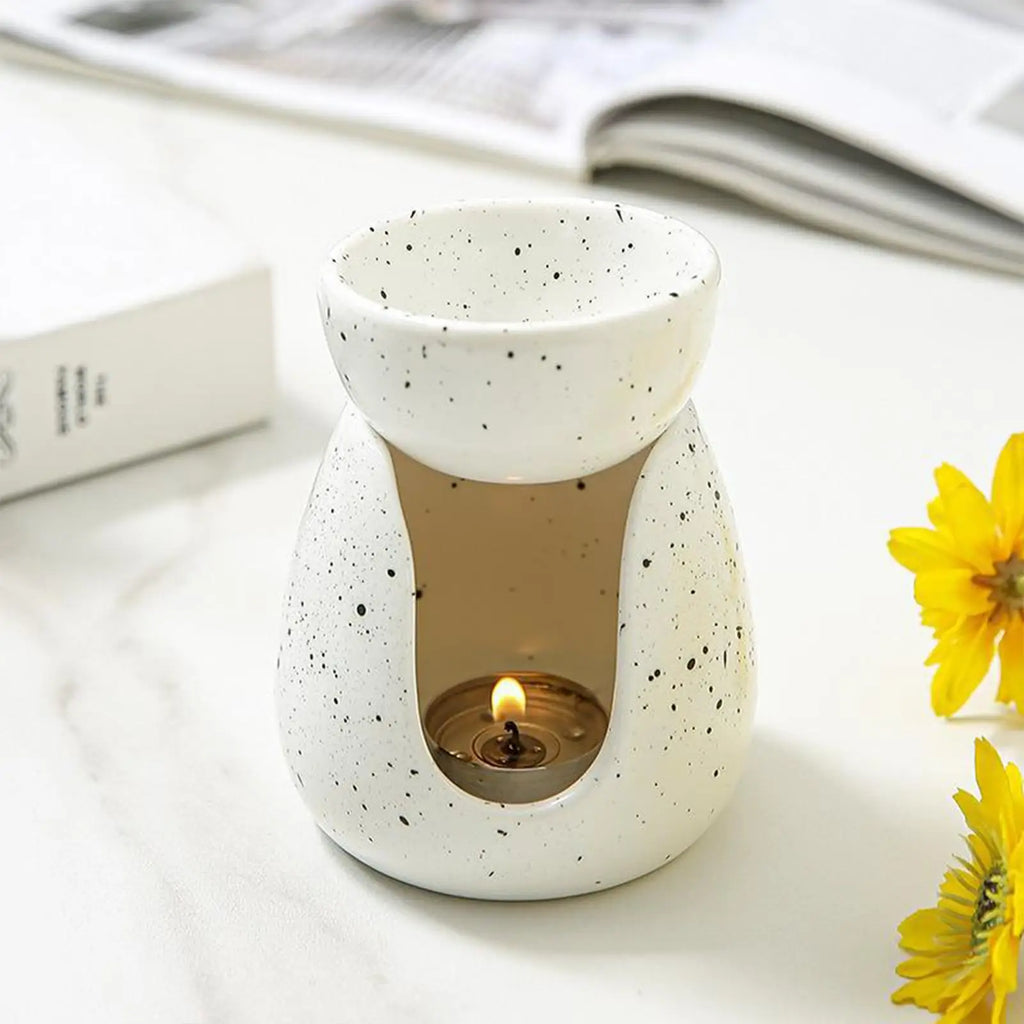 White ceramic oil burner with speckled finish on a tabletop, lit tealight, and yellow flowers nearby