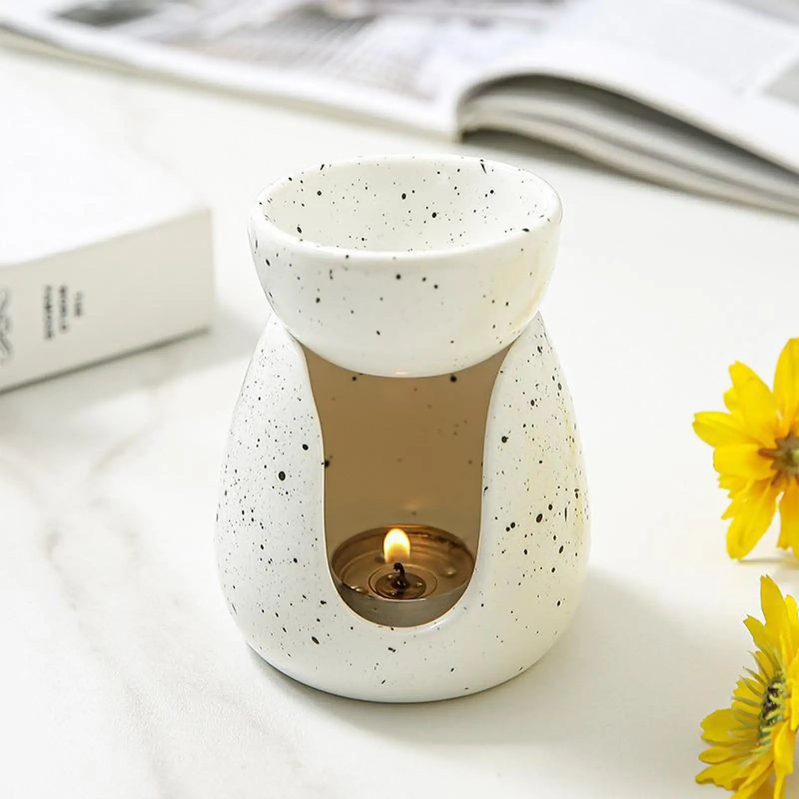 White ceramic oil burner with speckled finish on a tabletop, lit tealight, and yellow flowers nearby
