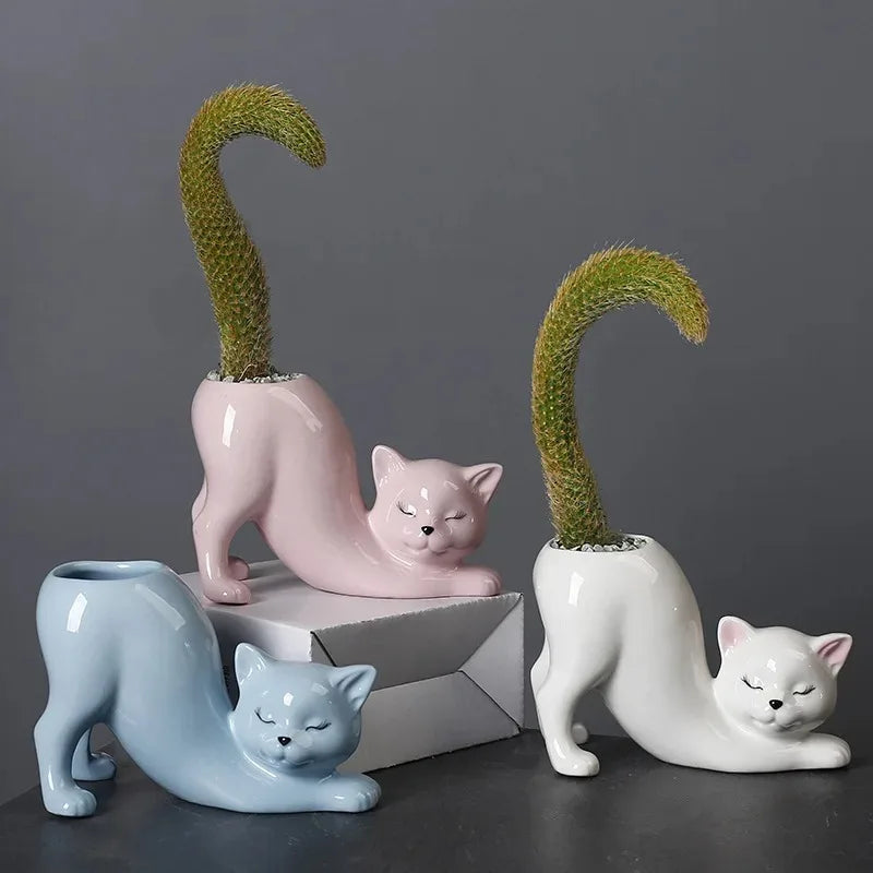 Three cat-shaped ceramic planters in white, pink, and blue, each holding a cactus, on a dark desk