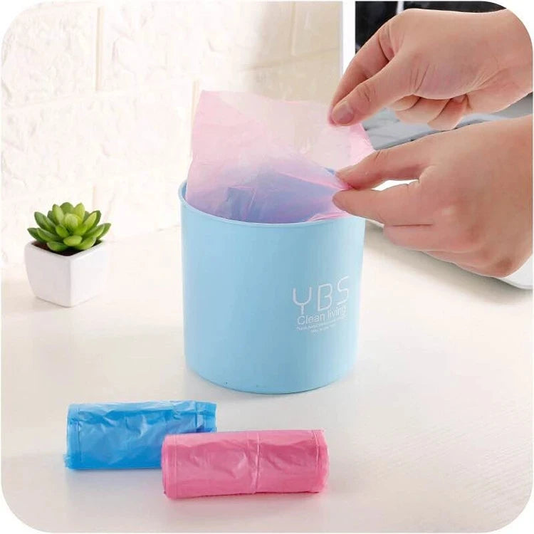 Hands placing a pink trash bag into a blue mini bin, with extra bag rolls on a table