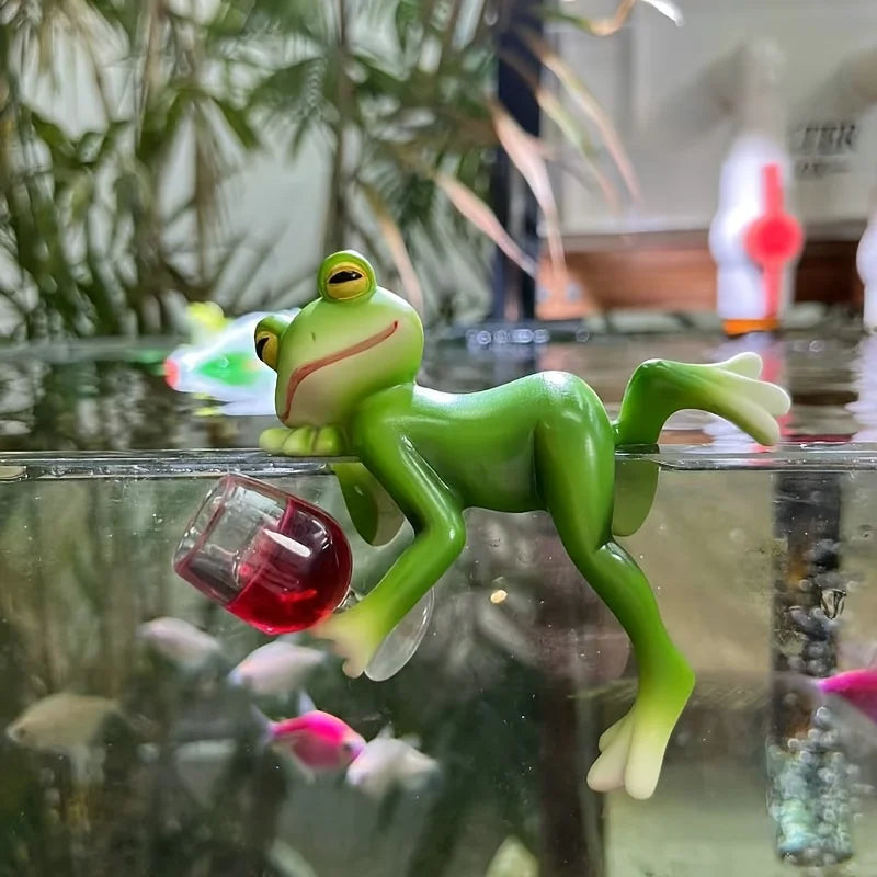Resin frog planter decoration posed with wine glass on the edge of a glass aquarium with fish and plants.
