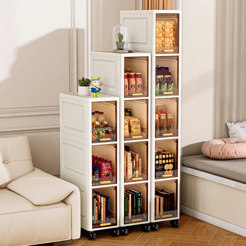 Three white slim rolling storage cabinets with multiple tiers, filled with snacks, books, and small containers beside a sofa and window seat