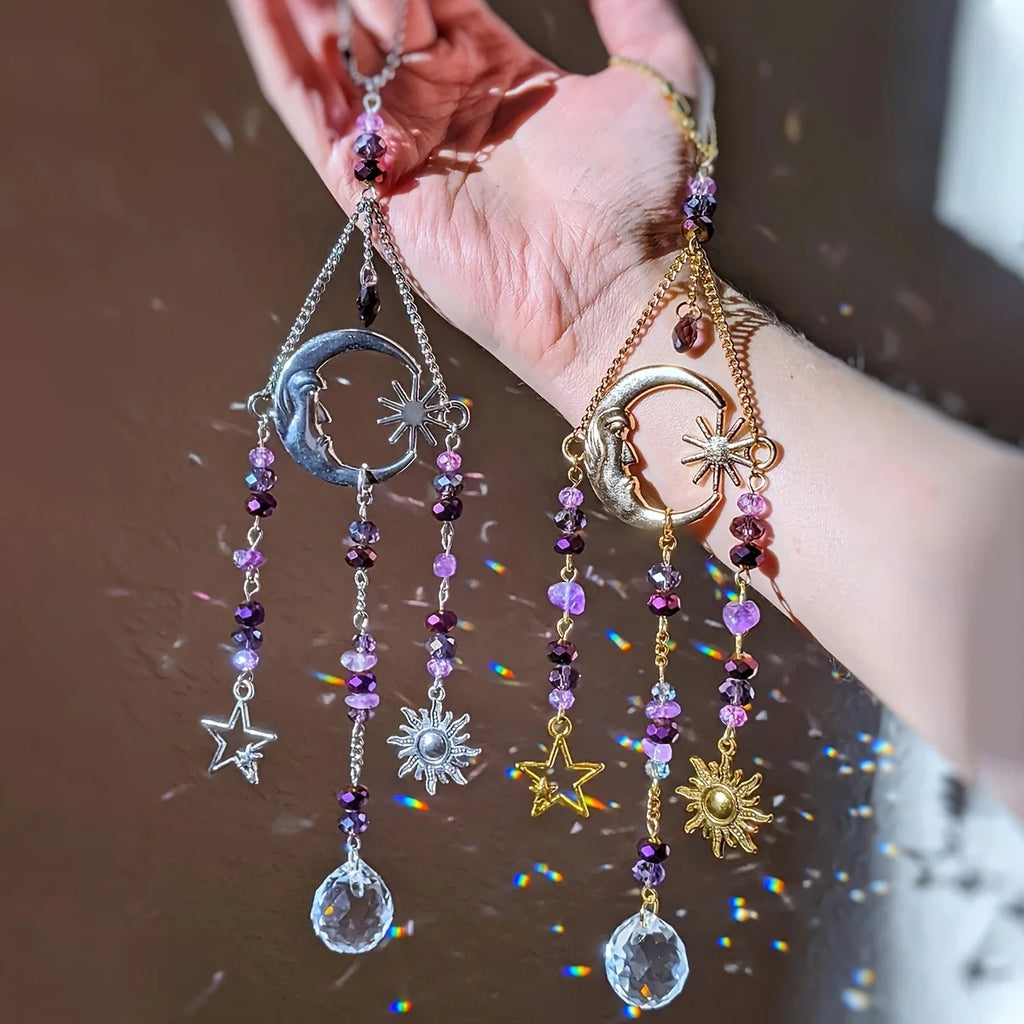 Two celestial crystal suncatchers with moon, sun, and star charms in silver and gold options held in a hand, catching rainbow light.