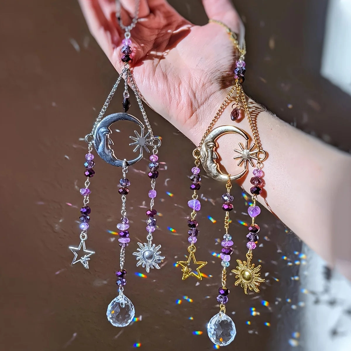 Two celestial crystal suncatchers with moon, sun, and star charms in silver and gold options held in a hand, catching rainbow light.
