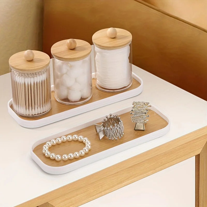 Slim white-edged vanity trays on a light wood surface holding glass jars with cotton, cotton swabs, and jewelry