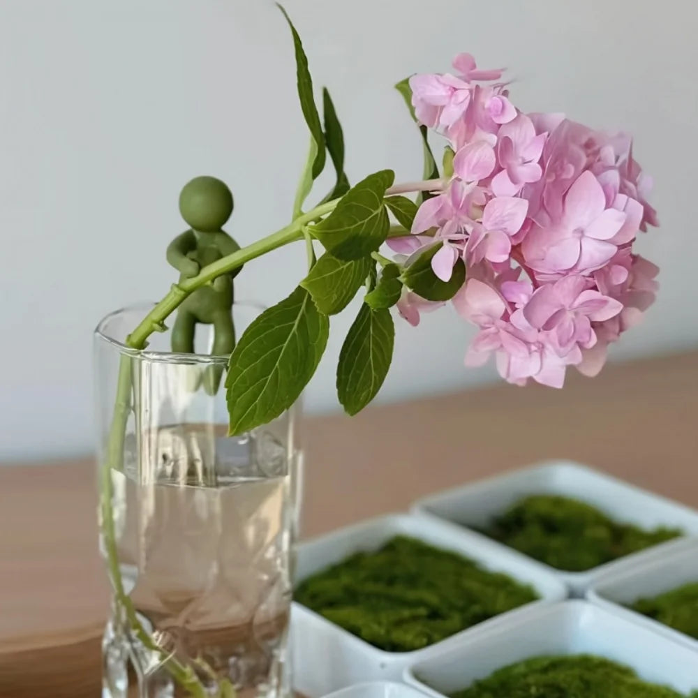 Green plant stem holder supporting a pink hydrangea stem in a glass vase next to small white dishes