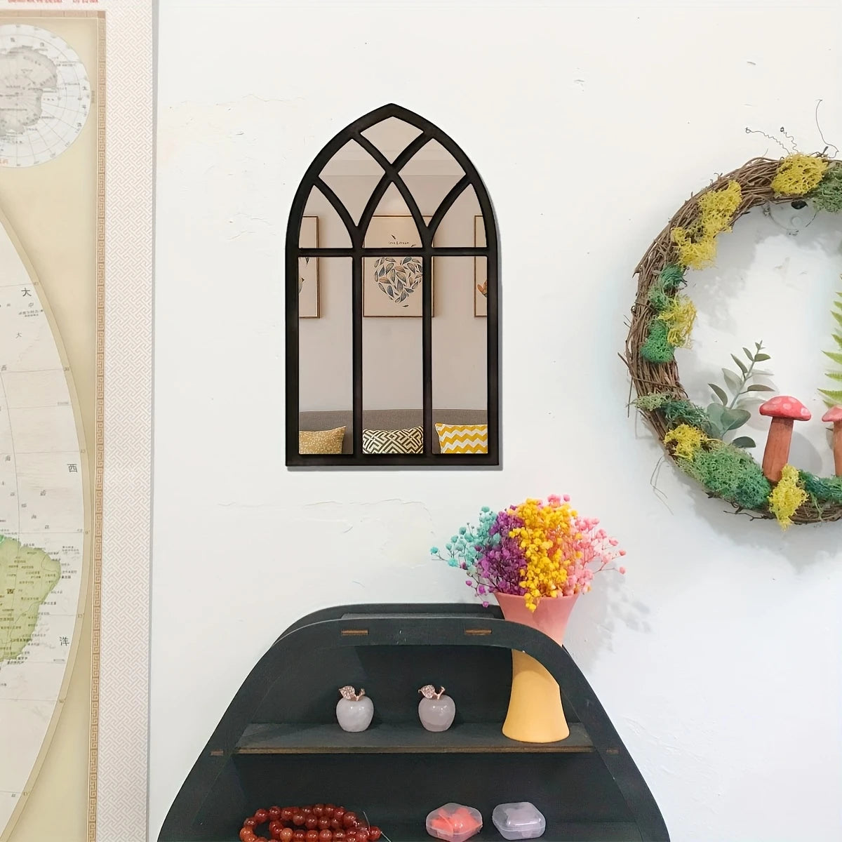 Gothic arch windowpane wall mirror with a black frame hanging on a white wall above a decorative shelf and wreath
