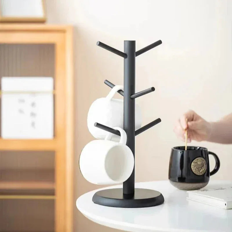 Black wooden tree-shaped mug holder with six hooks holding white mugs on round base, styled on modern kitchen table.