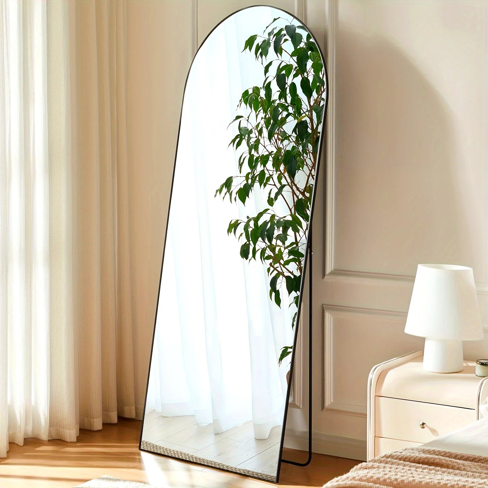 Arched full length mirror with slim black aluminum frame standing in a bright bedroom corner near a nightstand and leafy plant